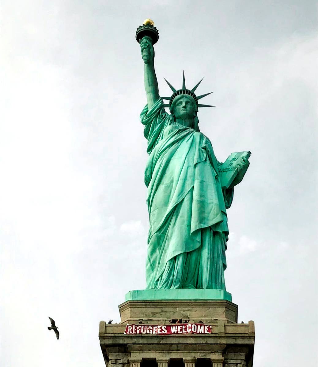 In this photo provided by Vitória Londero, a giant banner saying ‘Refugees Welcome’ hangs on the pedestal of the Statue of Liberty on Tuesday, Feb. 21, 2017, in New York. 