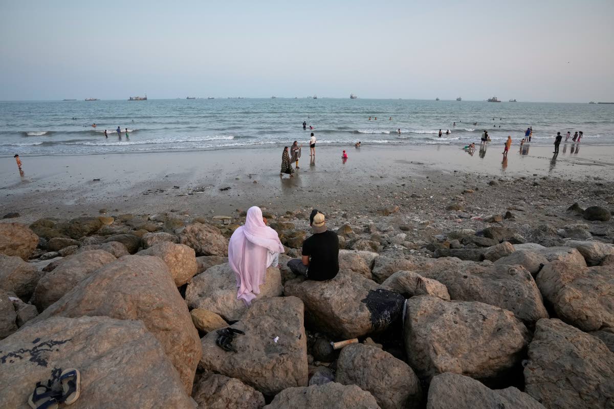 Two people sit on rocks overlooking a beach as others swim and commercial vessels lie anchored in the Strait of Hormuz off Bandar Abbas, Iran.