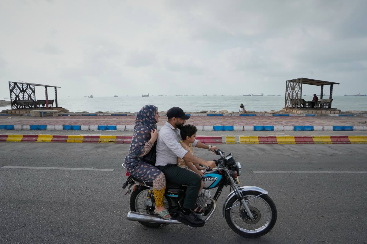 A man, woman and boy ride a motorcycle along the waterfront as commercial vessels lie anchored in the Strait of Hormuz off Bandar Abbas, Iran.